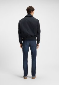 Muškarac odjeven u crnu bomber jaknu, tamnoplave traperice i sive cipele, stoji okrenut leđima na jednobojnoj bijeloj pozadini.