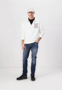 Fleece bianco con zip a quarti e logo "H" bordeaux, T-shirt nera sotto, jeans in denim blu e sneaker nere con suole grigie.