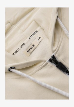 Beżowy kaptur z troczkiem, zamek błyskawiczny i miękka podszewka. Etykiety zawierają "Cotton Line" oraz rozmiar "M". Posiada fakturowaną powierzchnię materiału.