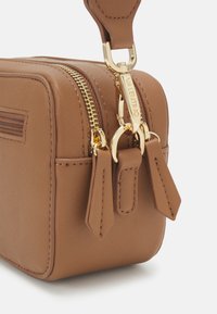 Bolsa de couro retangular em tom areia com ferragens douradas, apresentando um fecho com zíper e puxadores decorativos. Superfície texturizada com um brilho subtil.