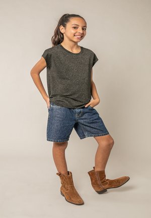 Fille aux cheveux foncés attachés en queue de cheval, portant un t-shirt noir, un short en jean et des bottes en daim marron, debout les mains dans les poches et un pied levé.