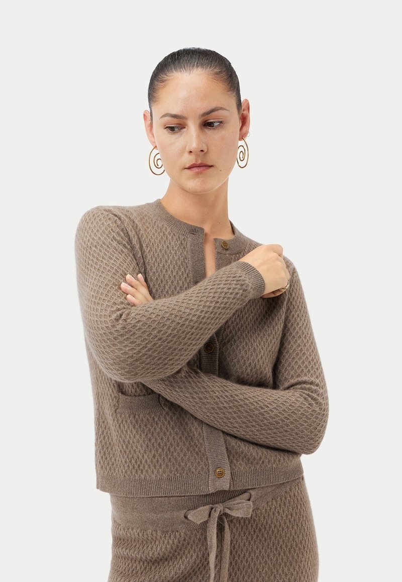 Brun stickad kofta med diamantmönster, rund halsringning och två knappar. Har långa ärmar och en kort passform med sidofickor.