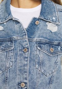 Light blue denim jacket with distressed details, metallic buttons, double chest pockets, and visible stitching accents.