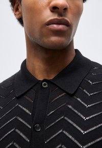 Primer plano de una persona que lleva una camisa negra con cuello, con patrones de malla en zigzag y dos botones visibles.