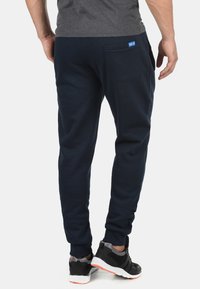 Navy jogger pants in soft fabric with elastic cuffs and a rear pocket. Features a blue logo tag near the pocket. Worn with black sneakers.