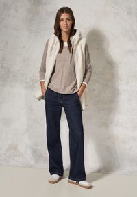 Gilet di crema chiaro senza maniche sopra un maglione di maglia taupe, abbinato a jeans blu scuro e sneakers bianche con suole marroni.