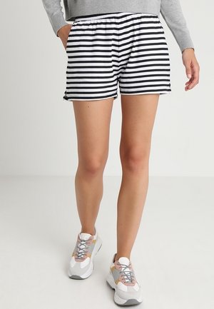 Person iført sorte og hvide vandrette stribede shorts, en grå sweater og multifarvede chunky sneakers står på en ensfarvet lys baggrund.