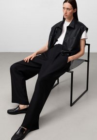 Giacca senza maniche in pelle nera sopra una maglietta bianca, abbinata a pantaloni neri larghi e mocassini neri. Design minimalista e moderno.