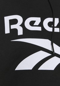 Czarna bluza z kapturem z dużym białym logo "Reebok" oraz zaokrąglonym elementem wzorniczym. Materiał wydaje się miękki z delikatną teksturą.