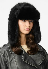 Czarna, sztuczna czapka trapper z szerokim, płaskim wierzchem, klapkami na uszy i miękką teksturą, noszona przez modela w skórzanej kurtce.