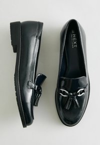 Schwarze Lackleder-Loafers mit abgerundeter Spitze, ausgestattet mit einer dekorativen Metallkette und Quasten auf dem Blatt, niedriger Blockabsatz für zusätzlichen Halt.