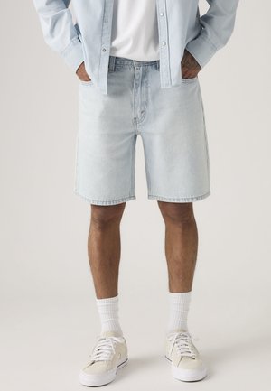 Inimene, kes kannab helesiniseid teksapükse, valgeid tossusid, valgeid sokke, valget t-särki ja helesinist teksast pikkade varrukatega pluusi, hoides käsi taskutes.