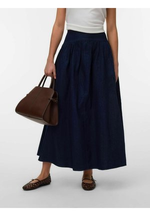 Femme portant une longue jupe bleu foncé et un haut blanc, tenant un sac à main marron et portant des ballerines tissées marron.