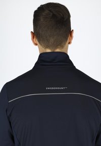 Marineblå jakke med høj krave, der har en reflekterende sølvkant langs skulderen og "SWEDEMOUNT™" logoet på ryggen.