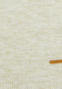 Écharpe en tricot couleur crème avec une texture douce et de légères variations d'ombre. Comprend une petite étiquette en bois portant le nom de la marque "BARTS."