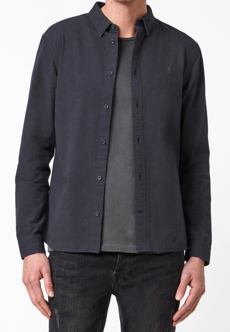 Tmavě modrá košile na knoflíky vyrobená z měkké látky. Má dlouhé rukávy, límeček a přední kapsu, kterou má položenou přes šedé tričko.