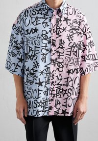 Camiseta de manga corta con un diseño dividido: una mitad azul claro con rayas y la otra rosa suave, ambas adornadas con texto en estilo graffiti negro y estrellas.