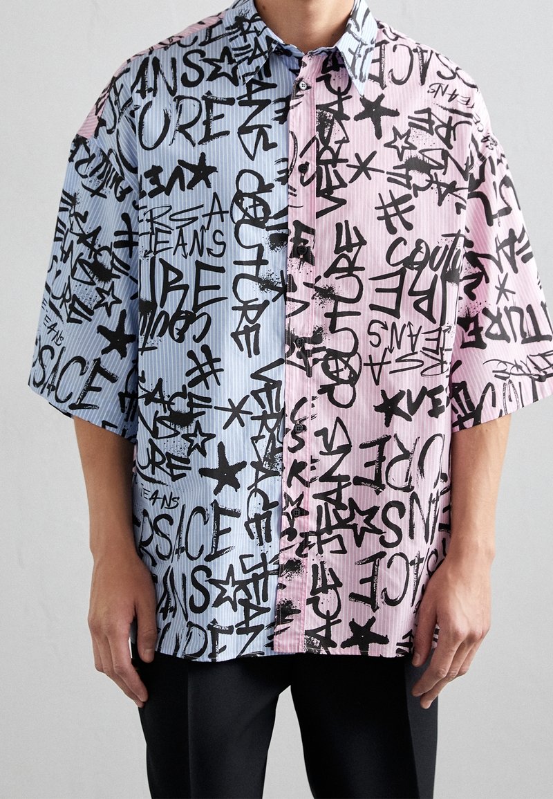 Camiseta de manga corta con un diseño dividido: una mitad azul claro con rayas y la otra rosa suave, ambas adornadas con texto en estilo graffiti negro y estrellas.