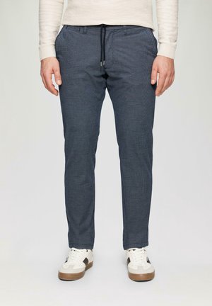 Mann trägt eng geschnittene dunkelblaue Hose mit Kordelzug und weiße Sneakers, steht vor einem schlichten hellen Hintergrund.