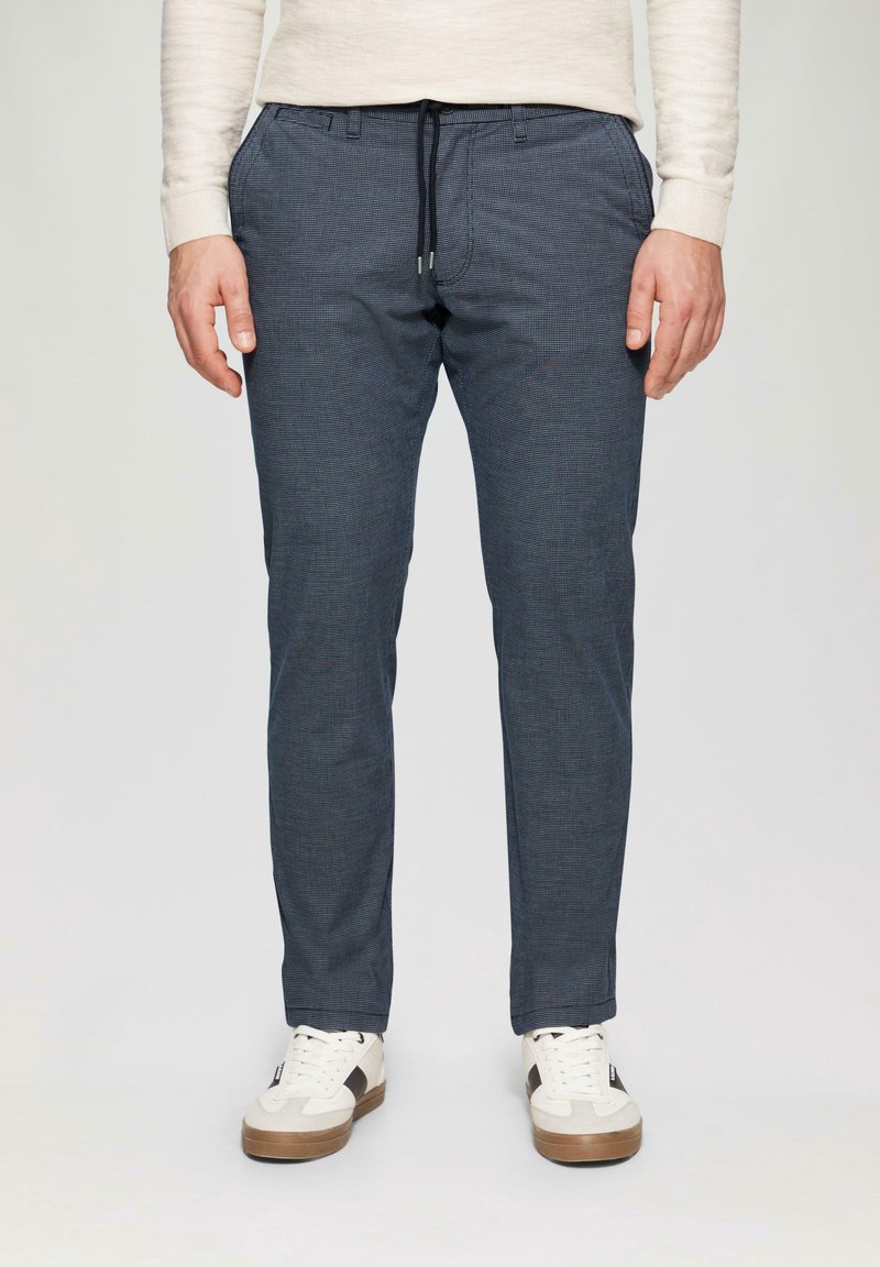 Homme portant un pantalon bleu marine slim à cordon et des baskets blanches, debout devant un fond clair uni.