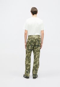 Kaimanu bikses zaļā toņos, ar brīvu piegriezumu, teksturētu audumu un gludu jostasvietu. Valkātas ar vienkāršu baltu T-kreklu.