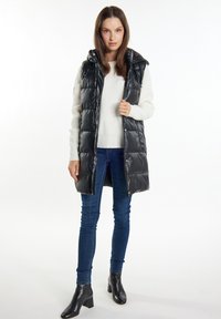 Sort polstret vest med hætte, der har en blank overflade, frontknapper og sidelommer, båret over en hvid sweater og blå jeans.