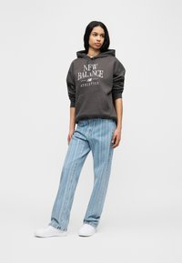 Γυναίκα όρθια με casual outfit, φοράει σκούρα γκρι φούτερ New Balance, ανοιχτού χρώματος τζιν με ρίγες και λευκά αθλητικά παπούτσια, σε απλό φόντο.