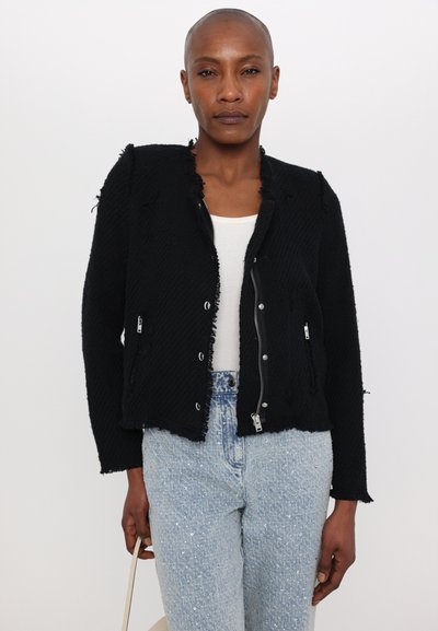 Schwarze cropped Jacke aus strukturiertem Stoff mit ausgefransten Kanten, vorderem Reißverschluss und Druckknöpfen, getragen über einem weißen Oberteil und hellblauen Jeans.