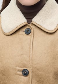 Κοντινό πλάνο μιας μπεζ παλτό από shearling με μαύρα κουμπιά και λευκή fleece γιακά που φοριέται πάνω από καφέ ζιβάγκο.