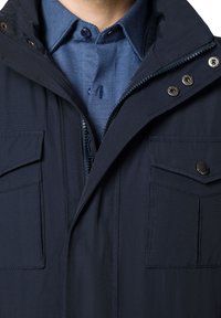 Granatowa kurtka z zapięciem na zamek i guzikami, z dwoma przednimi kieszeniami i wyłożonym kołnierzem na niebieskiej koszuli w wzory.