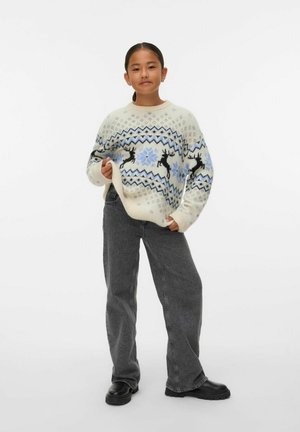 Sweter robiony na drutach w kremowym kolorze z niebieskimi i czarnymi wzorami reniferów oraz gwiazdek śnieżnych, w połączeniu z szarymi dżinsami o szerokiej nogawce i czarnymi butami.