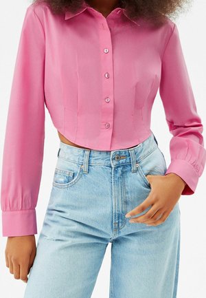 Woman wearing a fitted pink button-up crop blouse with long sleeves and light blue high-waisted denim jeans.