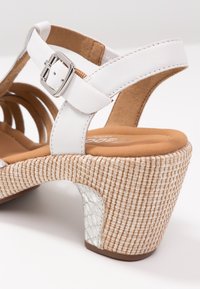 Białe sandały na paski z klamrą, z teksturowaną plecioną obcasem i miękką brązową wkładką. Uzupełnione błyszczącym białym akcentem na obcasie.