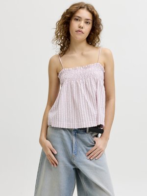 Jeune femme aux cheveux bouclés portant un haut sans manches à rayures roses et un jean bleu clair ample, debout les mains dans les poches.