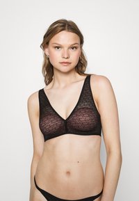 Svart bralette med et gjennomsiktig stoff som har et repetert mønster. Designet inkluderer en V-hals og brede stropper for støtte.