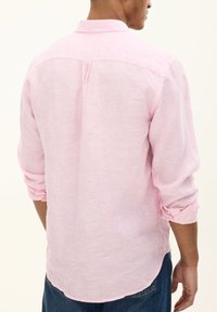 Camicia di lino rosa chiaro con maniche lunghe, colletto button-down, dettaglio della piega posteriore e polsini arrotolati, abbinata a jeans blu.
