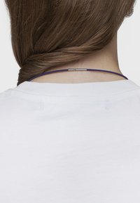 Collana sottile viola con design circolare liscio e chiusura metallica, indossata sul retro del collo, su uno sfondo di maglietta bianca.