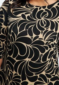 Gros plan d'un motif floral abstrait noir sur un tissu beige porté par une personne aux cheveux foncés, montrant le torse supérieur et une partie du bras.