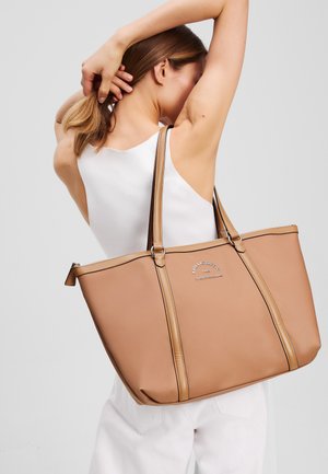 Grote beige tote bag van glad materiaal, met dubbele handvaten, een ritsluiting en contrasterende stiksels met reliëflogo-detail.