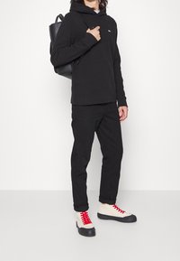 Sudadera negra de manga larga con capucha y un logo, combinada con pantalones negros y zapatillas color crema con cordones rojos y suelas negras.