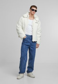 Puffatakki off-white värissä, jossa on korkeakaulus ja vetoketju edessä, yhdistettynä sinisiin suoriin farkkuihin ja valkoisiin lenkkareihin. Tekstuuriset materiaalit.