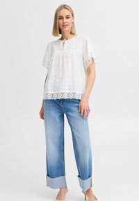 Blusa bianca a maniche corte con dettagli in pizzo e scollatura a chiave, abbinata a jeans a gamba larga blu chiaro con risvolti.