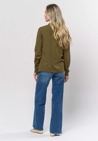 Oliivinvihreä pitkähihaisen suoralla leikkauksella ja pyöreällä kaula-aukolla, yhdistetty vaaleansinisiin leveälahkeisiin farkkuihin ja beigen värisiin punottuihin kenkiin.