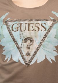 Devant d'un t-shirt marron avec un logo triangulaire clouté contenant "GUESS", un point d'interrogation, et un motif floral bleu clair en arrière-plan.