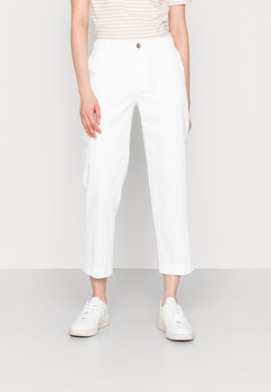 Kobieta w białych skróconych spodniach cargo, beżowo-białej koszulce w paski z krótkim rękawem oraz białych tenisówkach stoi na szarym podłożu.