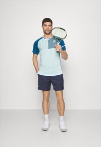 Camiseta deportiva azul claro con mangas azul marino y patrón de rayas verticales, combinada con pantalones cortos azul marino; sostiene una raqueta de tenis.