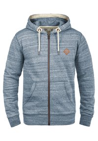 Sudadera con cremallera de color azul claro, hecha de una tela suave, que presenta un patrón texturizado, cordones y un acento de cuero en forma de diamante en el pecho.
