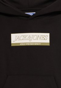 Hoodie noir avec un patch rectangulaire sur la poitrine portant les inscriptions "JACK & JONES" et "SINCE NINETEEN NINETY" en couleurs beige et olive.