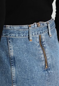 Gonna in denim con vita alta e chiusura con zip frontale. Presenta un tessuto blu con una texture sbiadita e cuciture in contrasto lungo le cuciture.