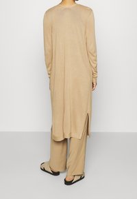 Beżowa tunika z długim rękawem o prostym kroju, z bocznymi rozcięciami i miękką fakturą; noszona na spodniach o szerokich nogawkach w tym samym odcieniu beżu oraz sandałach.